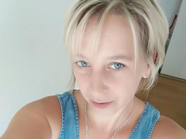 HeisseMarissa-hot has blonde hair, blue eyes, and is wearing a denim sleeveless top with a silver chain necklace. Her small tits are visible. She has a cute smile.
