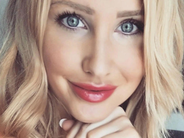 Fitness-Maus has blonde wavy hair, blue eyes, and a slight smile with pink lipstick. She's touching her chin with her hand, showing off her white nails.