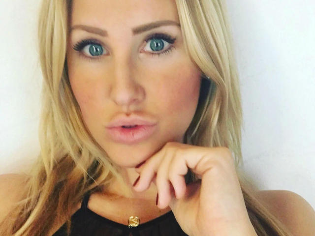 Fitness-Maus has blue eyes, blonde hair, and full pink lips, with her hand near her chin, wearing a black top with a gold button, looking at the camera.