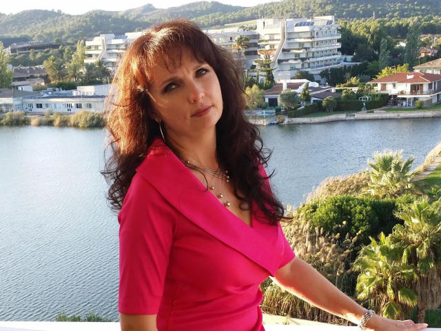 Hot-Lady34 with brown wavy hair, wearing a pink V-neck top, showing her small tits and necklace, standing by a waterfront with palm trees and mountains in the background.