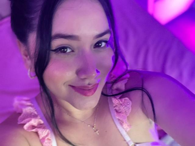 VeronicaWins with pink ruffled top, dark hair in ponytail, nose ring, smiling, purple lighting, earrings, necklace, subtle makeup, pink lips, cute, sexy, soft light, intimate selfie.