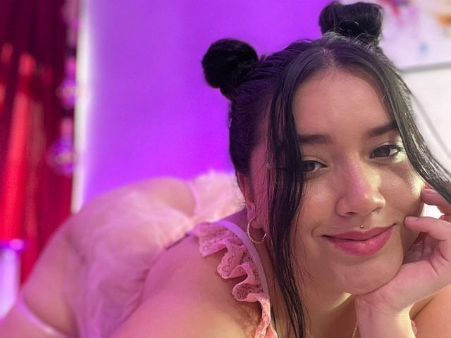 VeronicaWins with black double buns, wearing pink lace lingerie, smiling at the camera, showing a nose ring and touching her cheek, with her ass and pink lace lingerie visible.