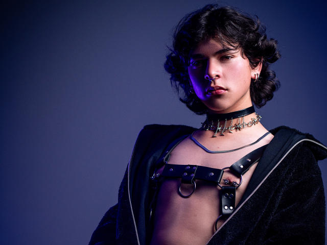 LukeHowlandy with curly dark hair, wearing a black leather harness, choker with chain details, and a black jacket, looking serious, with purple lighting on their face.