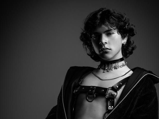 Luke Howlandy, avec ses cheveux bouclés, portant un harnais en cuir noir, un collier avec des pointes et une veste en velours sombre, a l'air sérieux sur une photo en noir et blanc. Les seins sont couverts, aucune fesse n'est visible..