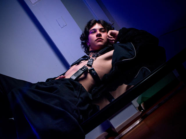 LukeHowlandy with wavy black hair, purple eyeshadow, black leather harness, and black pants, sitting on a table, looking down with a slight smirk, in a dimly lit room.