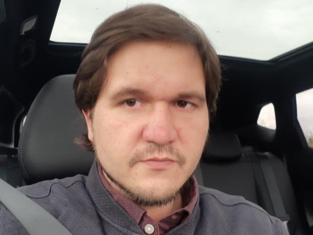 NectarineFad, a chubby guy with brown hair, a mustache, and a goatee, sitting in a car, wearing a gray shirt and a red button-up underneath.