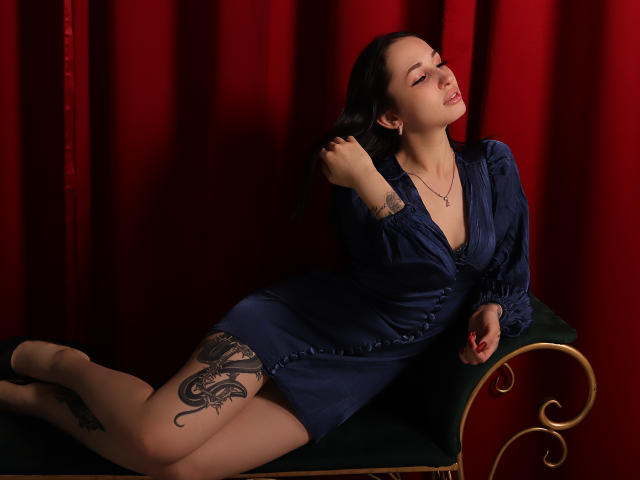 NatalyOlsen with long black hair, blue dress, tattoo on thigh, lying on green and gold couch, red curtain, hand in hair, eye closed, necklace, red nails, seductive pose.