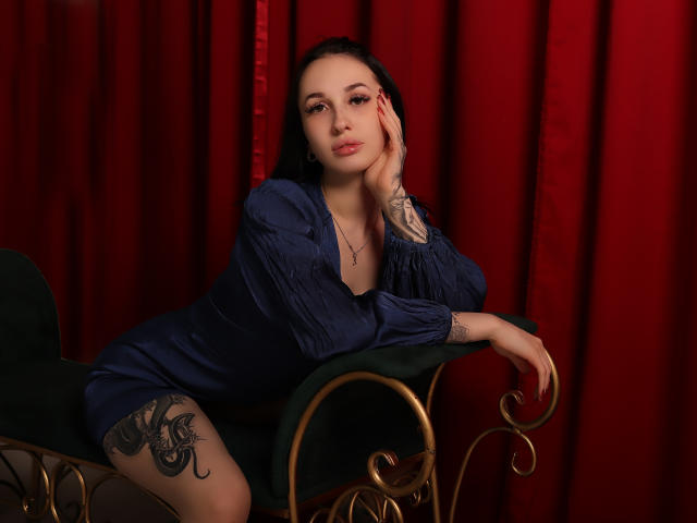 NatalyOlsen with dark hair, blue dress, and tattoos, leaning on a green chair, red curtains, hand on cheek, looking at camera, showing her tits and ass.