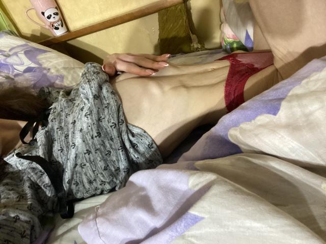 Kabblasha's slim, pale, flat-stomached body lies on a bed, wearing red lace panties, with a gray patterned shirt partially covering her, and her hand resting on her side.