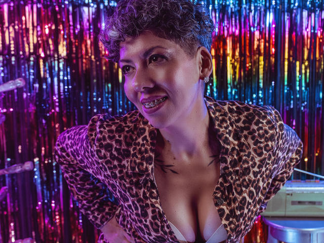 JuliaNewell with short curly hair, wearing a leopard print top, showing her tits, smiling, colorful glitter background, tongue piercing, tattoos on chest, looking sexy, confident, and playful.