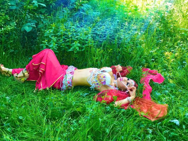 Catlady-hot lies on grass, wearing a red skirt, white floral top, and gold jewelry, with red sheer fabric, her hands near her mouth, in a lush green field.