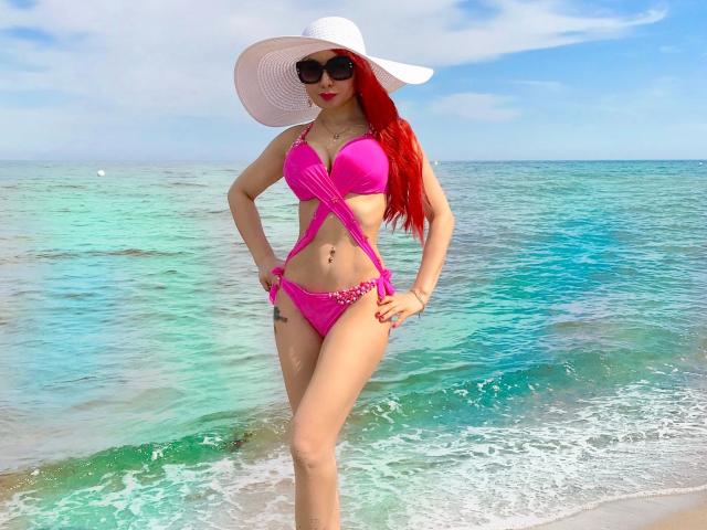 Catlady-hot with bright pink bikini, white sunhat, red hair, and black sunglasses, standing in clear turquoise water, showing her tits and ass, hands on hips, water at knees.