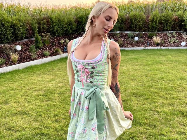 HannaSecret-hot with blonde hair, tattoos on right arm, wearing a green floral dress with pink ribbon, standing on grass, eyes closed, dress lifted slightly, showing her ass.