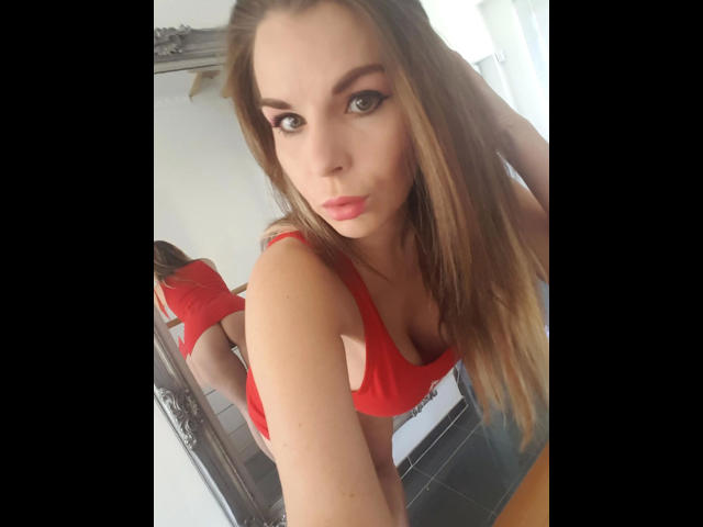 Aische-Pervers with long brown hair, red tank top, and red thong, bending over, showing her ass, and looking at the camera with pink lips and dark eyeliner.