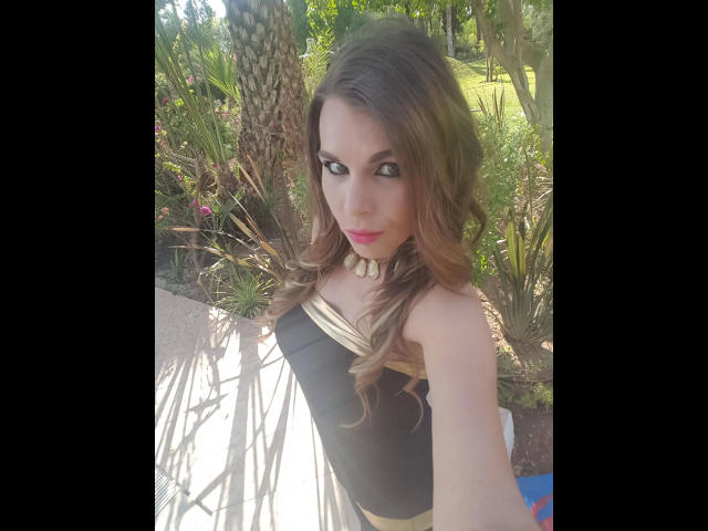 Aische-Pervers with long brown wavy hair, wearing a black and gold strapless dress, necklace with large beads, and pink lipstick, taking a selfie outdoors.