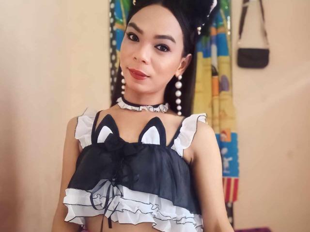 SugarVenom with dark hair in a bun, wearing a black and white frilled crop top with cat ears, a pearl choker, and pearl earrings, smiling at the camera.