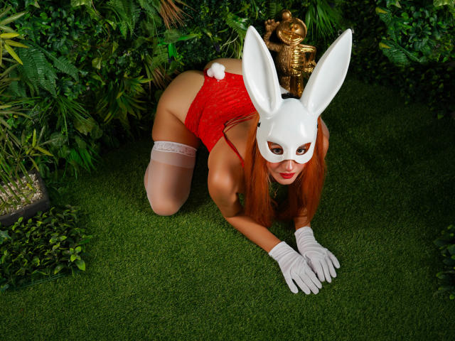 ChanneBell with red lingerie, white bunny mask, white gloves, and thigh-high stockings, on all fours, showing her ass, with a fluffy white bunny tail, on green grass.