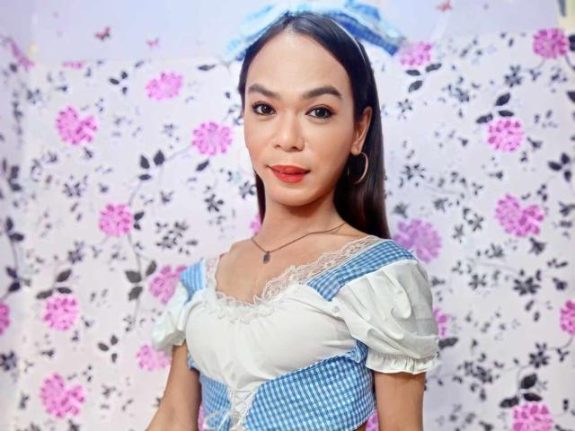 SugarVenom has long dark hair, wearing a blue and white checkered dress with lace, hoop earrings, and a blue headband, smiling with red lipstick, and a small necklace.