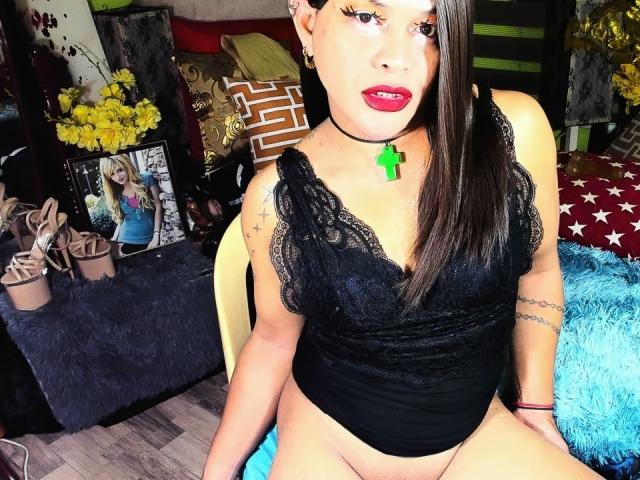 TransGiftedCum with long black hair, red lipstick, lace black top, green cross necklace, and exposed pussy, sitting on chair, blue fur pillow in background, yellow flowers on left.