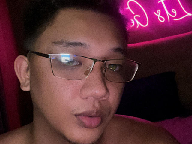 RoyalCum hat ein süßes Gesicht mit Brille, einem Nasenring und einem leichten Schnurrbart und blickt mit leicht geschürzten Lippen in die Kamera. Im Hintergrund ist ein neonpinkes Licht zu sehen..