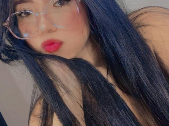 BellaDawson with long black hair, wearing clear glasses and red lipstick, pouting her full, red lips. Her bare shoulders are visible, focusing on her face.