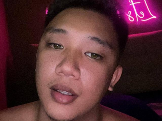 RoyalCum with short black hair, light brown skin, and green eyes, slightly open mouth with a small mustache, shirtless, looking into the camera. Neon pink light in the background.