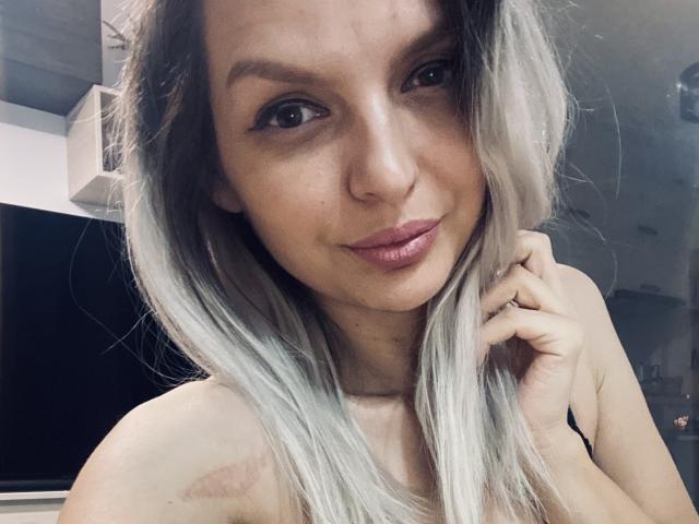 DouceAmelia with light blonde, wavy hair, smiling, looking at camera, hand touching her hair, slightly bruised shoulder, pink lipstick, brown eyes, light skin, subtle makeup, bare shoulders.