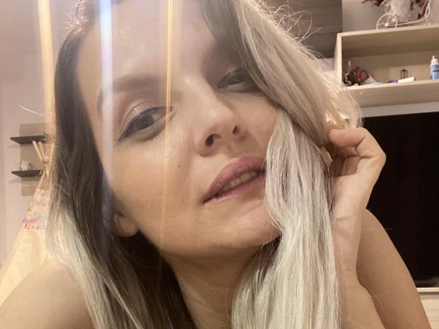 DouceAmelia with blonde hair, slightly parted lips, and a subtle smile, holding her hair, showing her nose and upper teeth, with a reflection in the upper left.