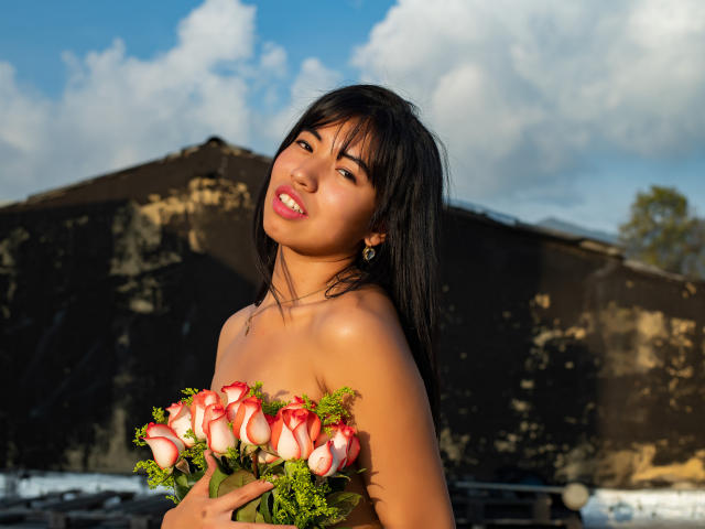 LuciaGarcia with black hair, holding a bouquet of pink and red roses, has tan skin and is smiling with pink lipstick, wearing a strapless top, and small earrings.