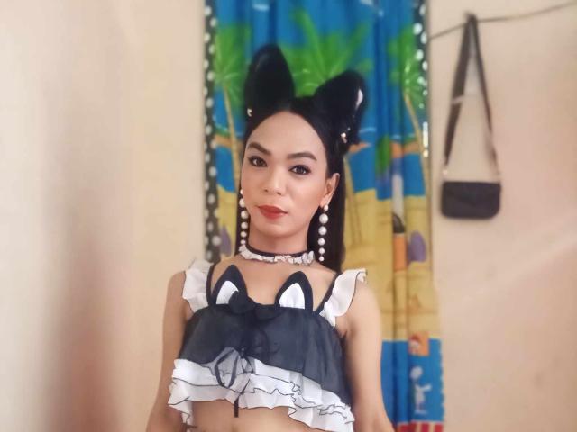SugarVenom with cat ears, wearing a black and white frilled top, showing her tits, adorned with pearl earrings and a choker, standing in a room with colorful curtains.