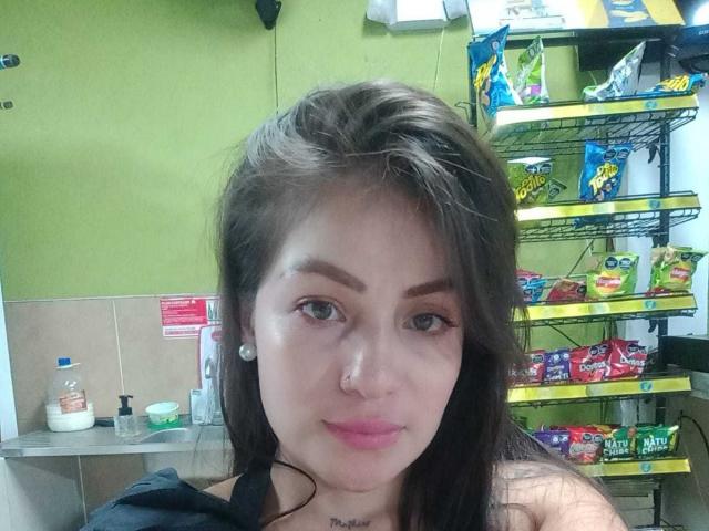 AngelaCutest has brown hair, green eyes, a nose ring, and pink lips, with small tits and a cute face, in a store with shelves of snacks in the background.