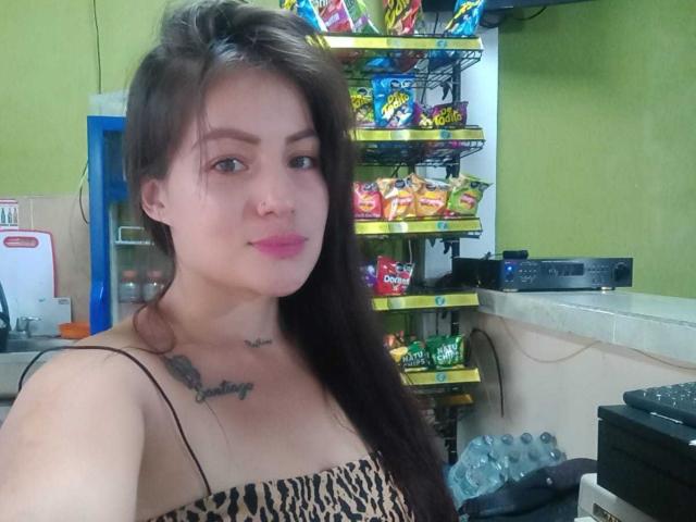 Angela: Eine Frau mit langen, braunen Haaren, rosa Lippenstift, kleinen Tattoos unter dem Kinn und auf der Brust, die ein Top mit Leopardenmuster trägt und in einem Laden steht, wobei im Hintergrund Snacks zu sehen sind..