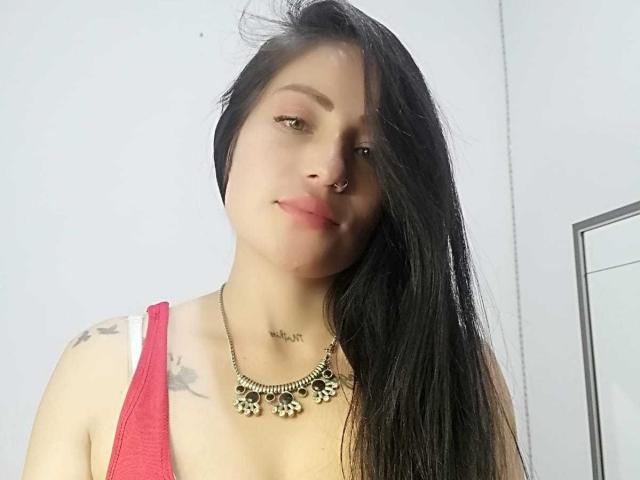 Angela: Cabelos pretos longos, piercing no nariz, top vermelho, colar com pingentes em forma de flores, pequenas tatuagens no ombro, sorriso discreto, pele clara e decote visível..