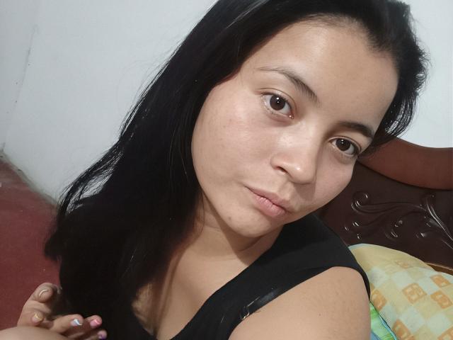 LiliHotter has puffy lips, brown eyes, and long black hair. She's wearing a black top, and her small tits are slightly visible. She's making a cute, pouty face.