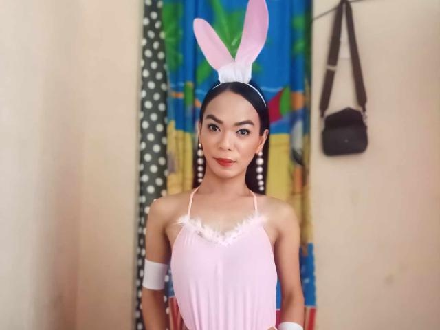 SugarVenom in pink lingerie, white arm bands, and pink bunny ears, standing with a cute smile, small tits, and a slim build, in a brightly colored room.