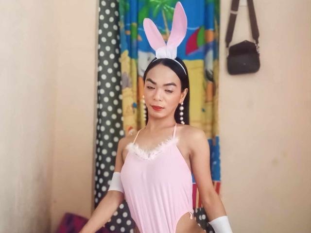 SugarVenom in a pink, cut-out, one-piece outfit with white fur trim, white gloves, and pink bunny ears, standing with a slight smile, looking down.
