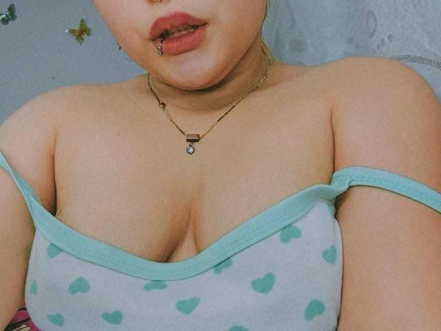 ElinaX with her tits showing, wearing a grey top with green hearts, off-shoulder, gold necklace, lip piercing, and light skin. Her chest is prominent.