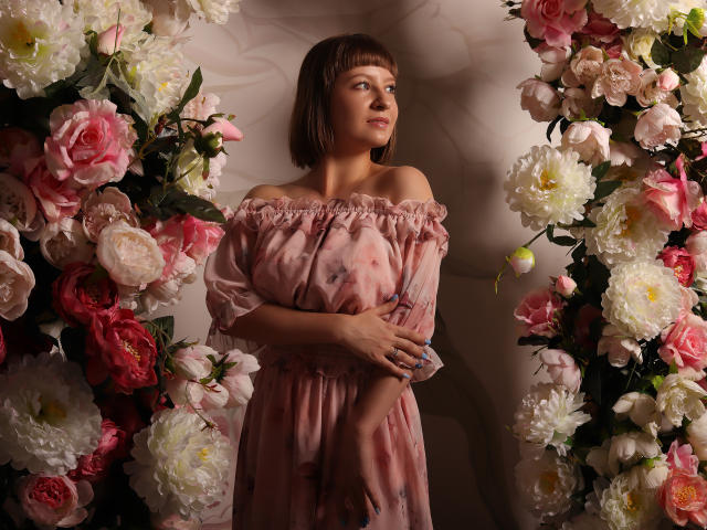 LizzyWest with short brown hair, off-shoulder pink floral dress, blue nails, standing between two tall floral arrangements, looking to the side. She has small tits and a cute face.
