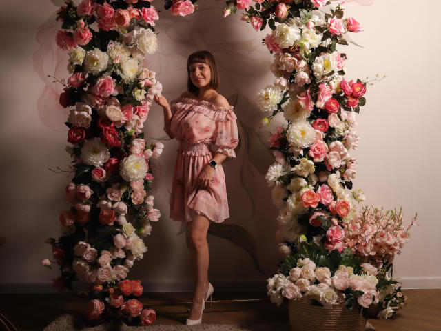 LizzyWest stands in front of a floral arch, wearing a pink off-shoulder dress with puffed sleeves, and white heels, surrounded by pink, white, and red flowers.