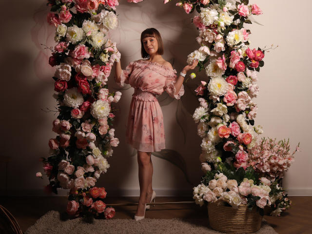 LizzyWest standing in front of a floral arch, wearing a pink floral dress, off-shoulder with puffy sleeves, white heels, holding flowers, surrounded by pink and white roses.