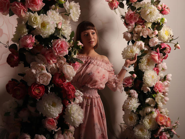 LizzyWest stands in front of a floral arch, wearing a pink, off-shoulder dress, holding a flower. She has short brown hair, a cute face, and small tits.
