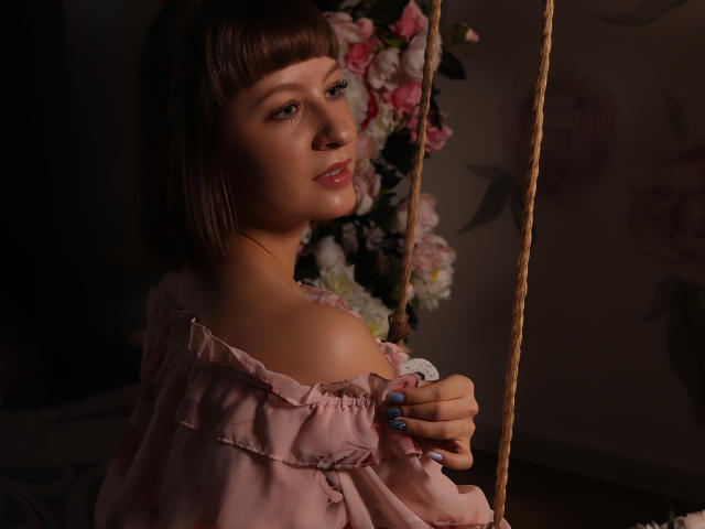LizzyWest with short brown hair, pink off-shoulder top, blue nails, sitting on a rope swing, looking over shoulder, flowers in background, soft lighting, subtle smile, gentle expression.