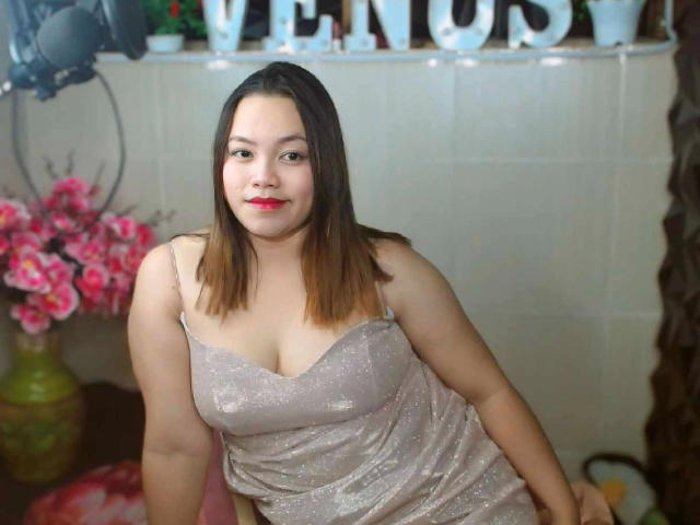 BedLover69-hot with medium brown skin, straight brown hair, wearing a sparkly pink dress, showing her boobs, sitting, smiling, red lipstick, pink flowers in the background.