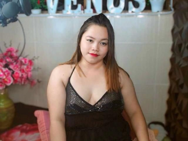 BedLover69-hot, chubby Asian woman with medium tits, wearing a black, sheer, sparkly dress, red lipstick, and straight brown hair, sitting, looking at camera, indoors, smiling.