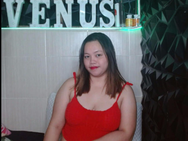 BedLover69-hot, a chubby Asian woman with red lipstick, wearing a red crop top, has medium tits and brown hair, sitting and smiling. Her red top is tied at the shoulders.
