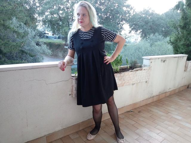 CuteDebby-hot stands with hands on hip, wearing black dress, striped shirt, black tights, and white flats, leaning on a cracked white wall, with blonde hair and red lipstick.