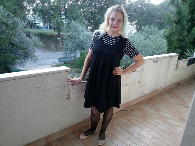 CuteDebby-hot with blonde hair, striped shirt, black dress, sheer black stockings, and silver flats, standing on tiled balcony, hand on wall, looking at camera.