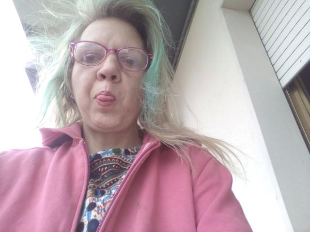 CuteDebby-hot with puffy pink jacket, colorful patterned shirt, and pink glasses, sticking out her tongue. She has blonde hair with green highlights, and a surprised expression.