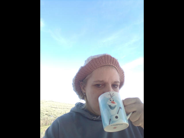 CuteDebby-hot with a red knit beanie, gray hoodie, holding a blue mug with a snowman design, sipping, under a bright blue sky.