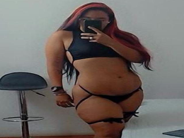 MiaTaylior, avec de longs cheveux roux et noirs, portant de la lingerie noire avec des bretelles, tenant un téléphone, debout devant un miroir, montrant ses seins et ses parties intimes..